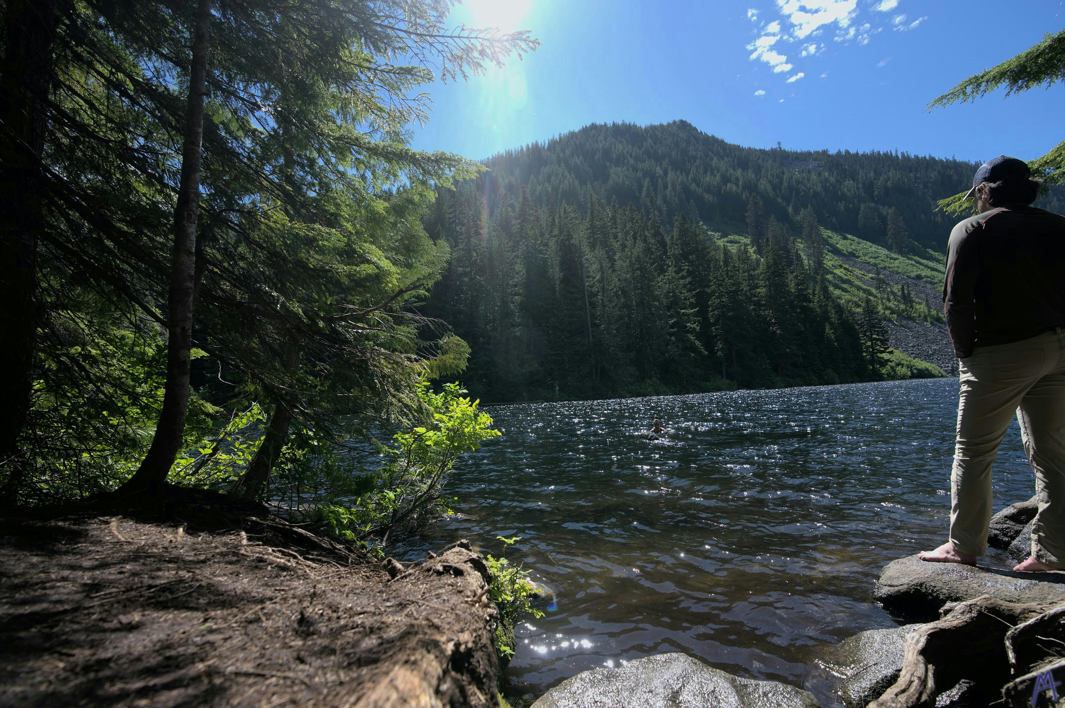 A man standing on a rock next to a clear lake with someone swimming in the mountains